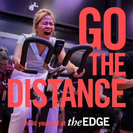 People cycling on exercise bikes. Girl in forefront has a determined expression. Text says: 'Go the distance. Build yourself and The Edge'